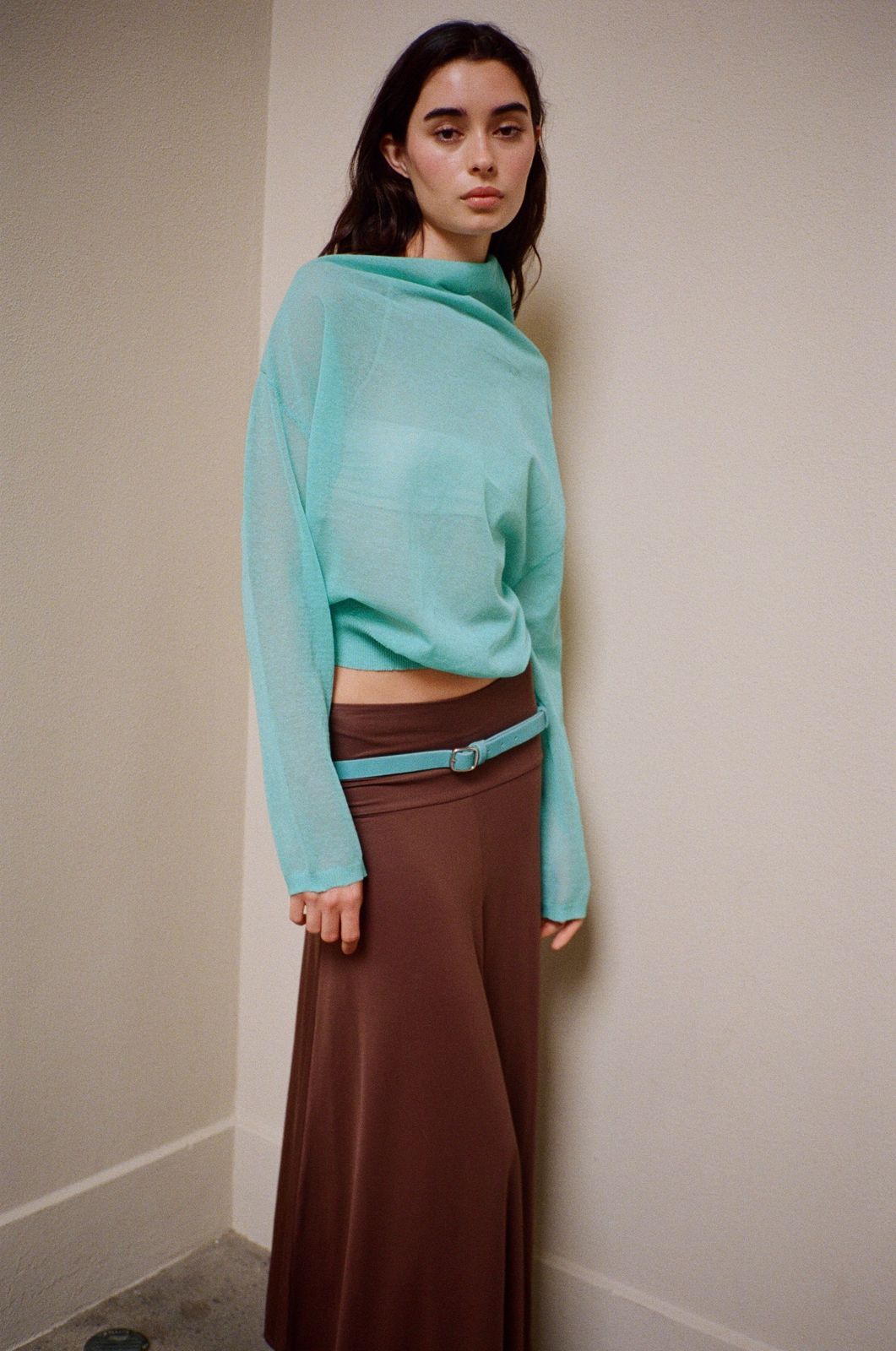 Woman wearing an aqua blue sheer sweater and brown pants on a beige background.