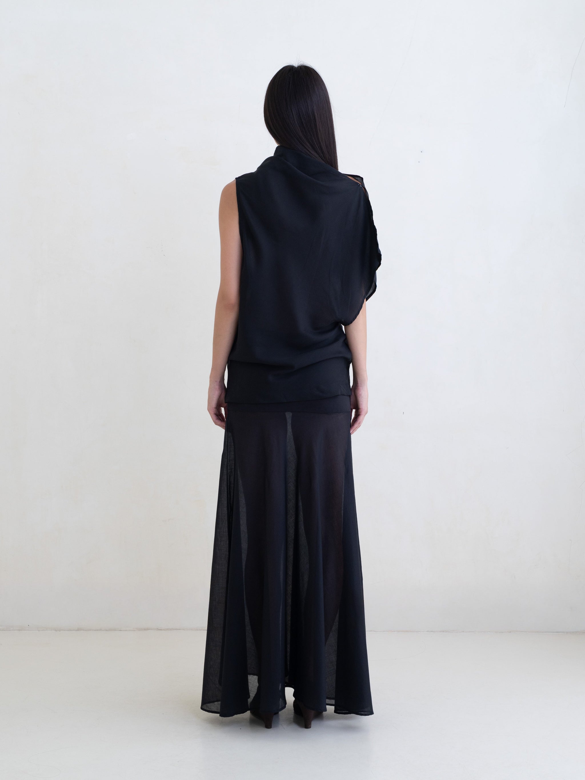 Woman wearing a black draped one-shoulder top and a black maxi skirt on a white background. 