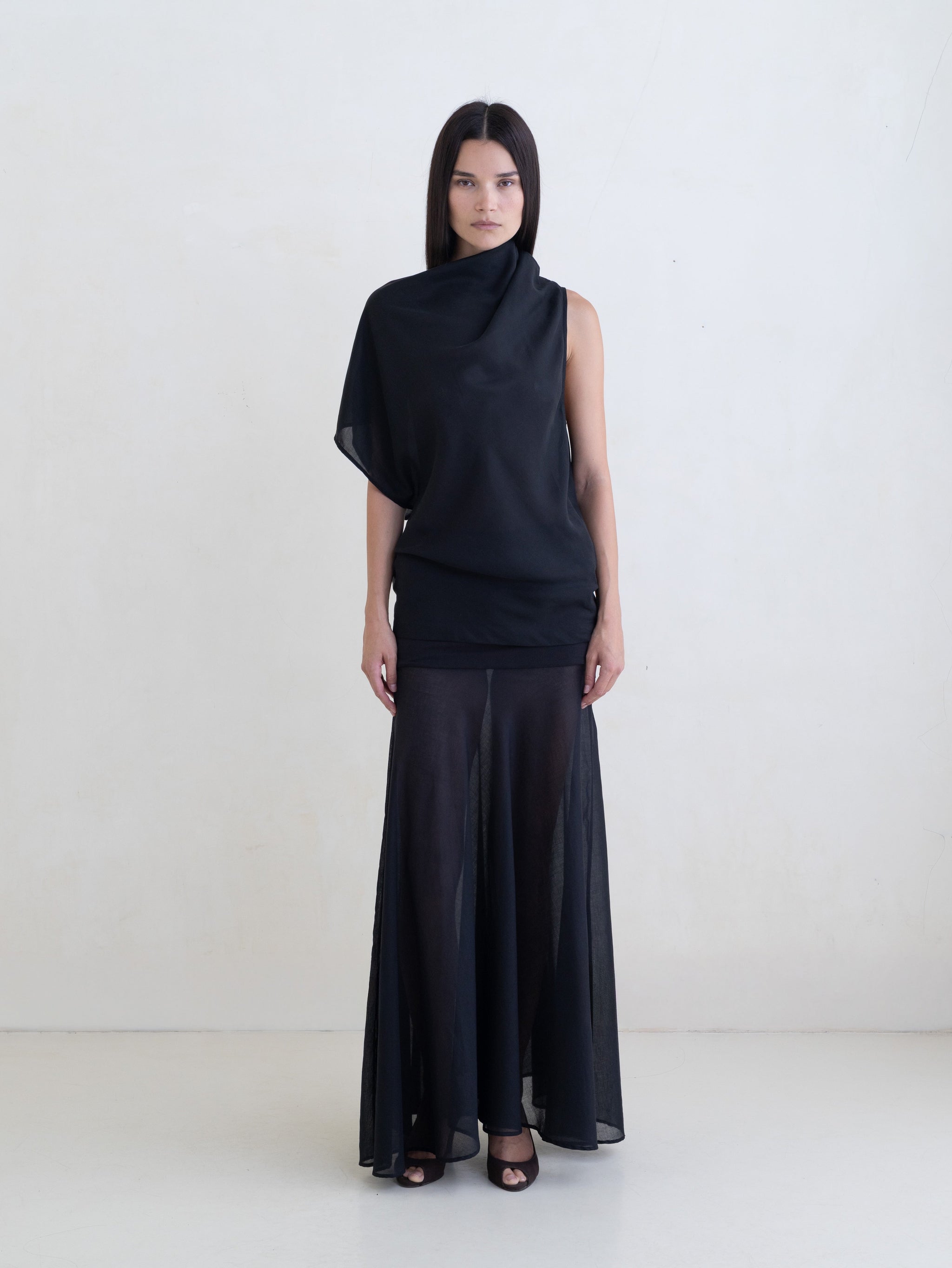 Woman wearing a black draped one-shoulder top and a black maxi skirt on a white background. 