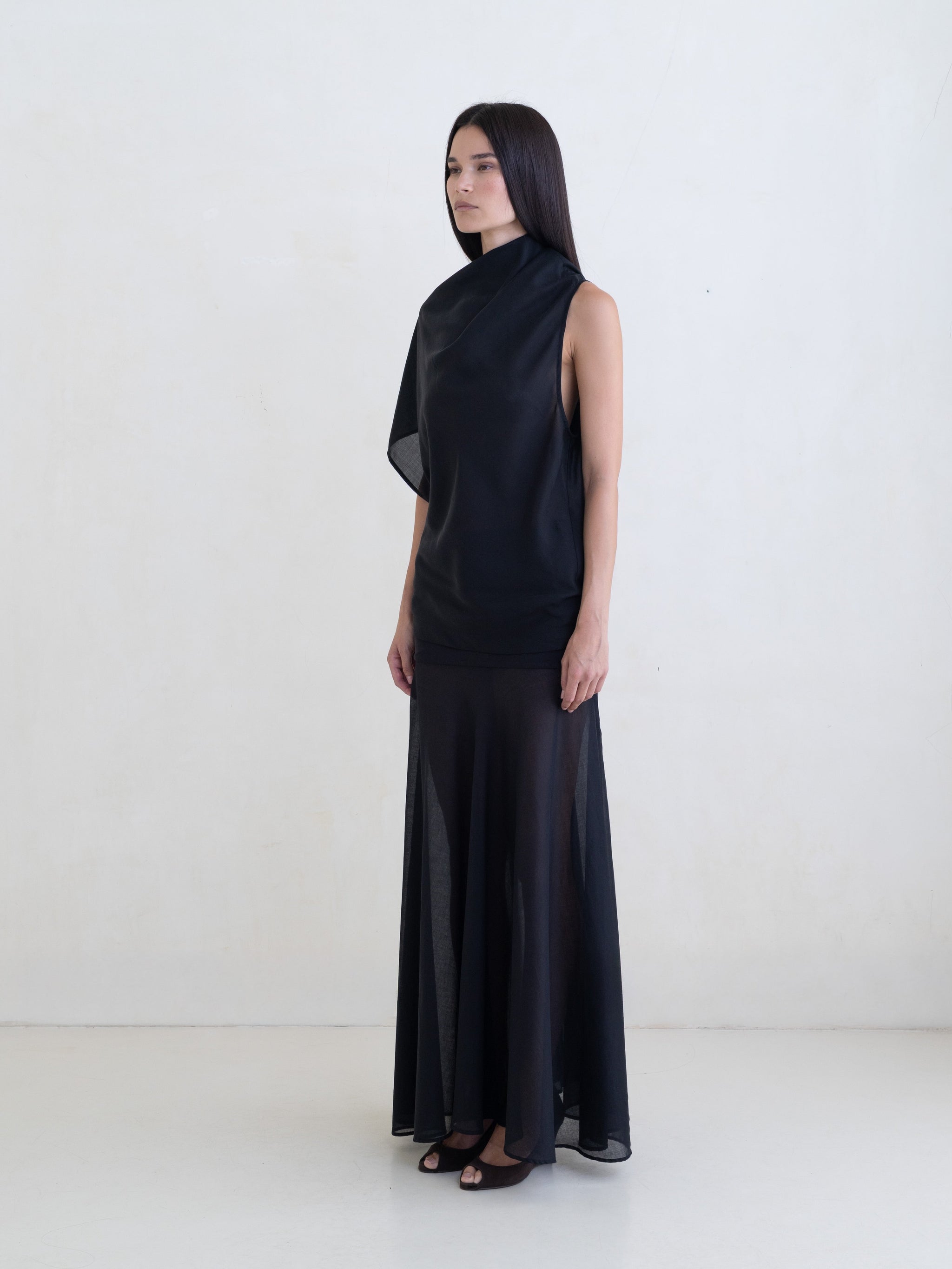 Woman wearing a black draped one-shoulder top and a black maxi skirt on a white background. 