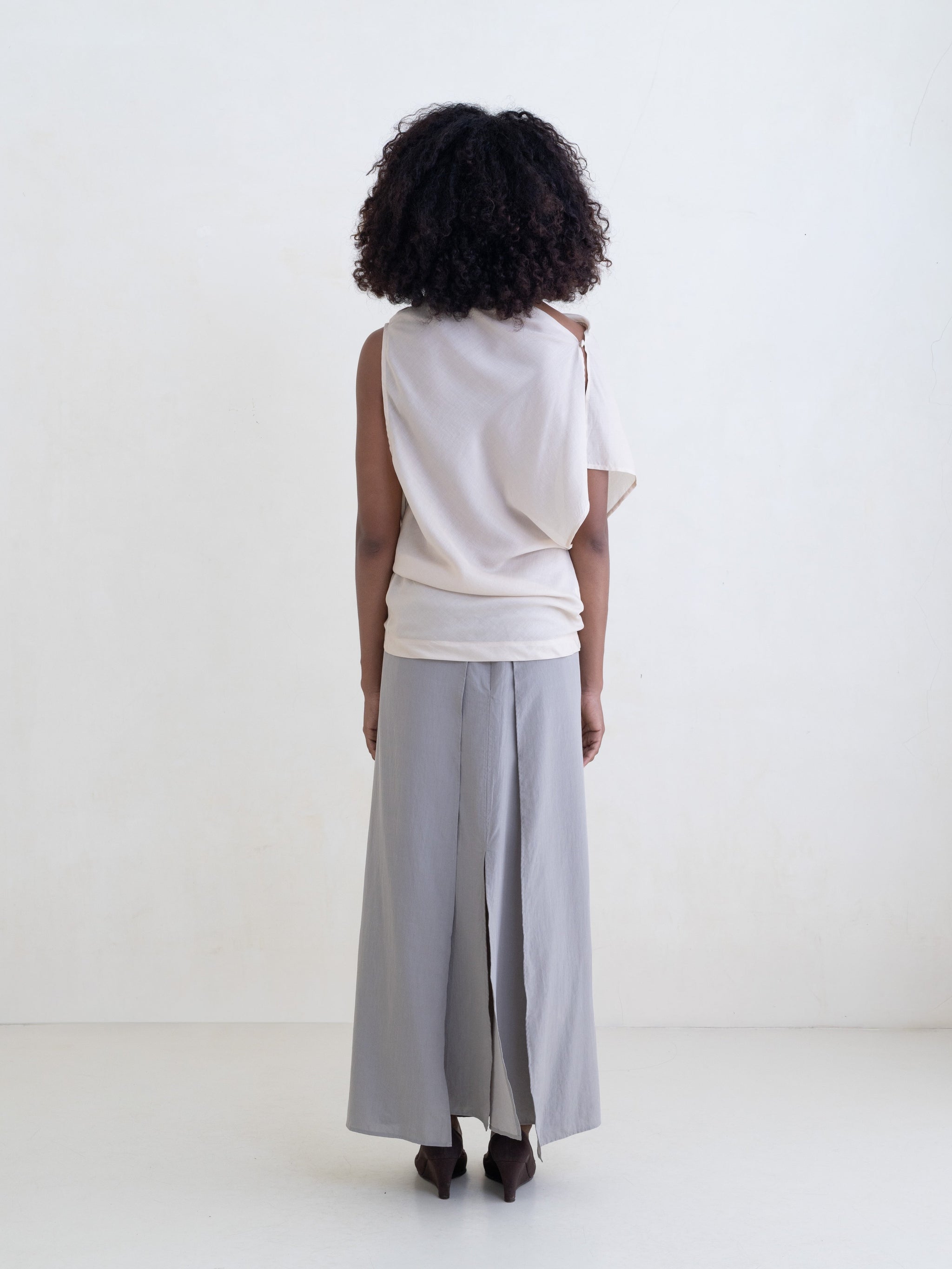 Woman wearing a cream draped one-shoulder top and a gray maxi skirt on a white background. 