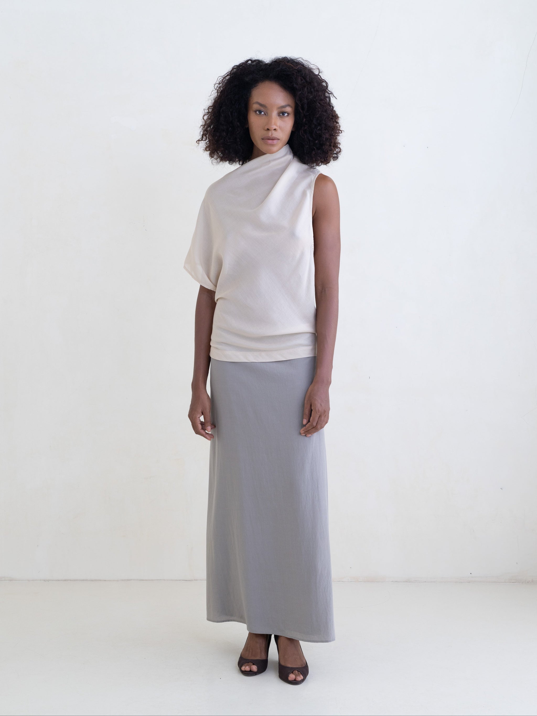 Woman wearing a cream draped one-shoulder top and a gray maxi skirt on a white background. 