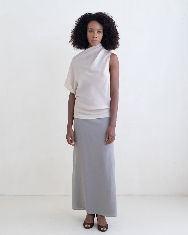 Woman wearing a cream draped one-shoulder top and a gray maxi skirt on a white background. 