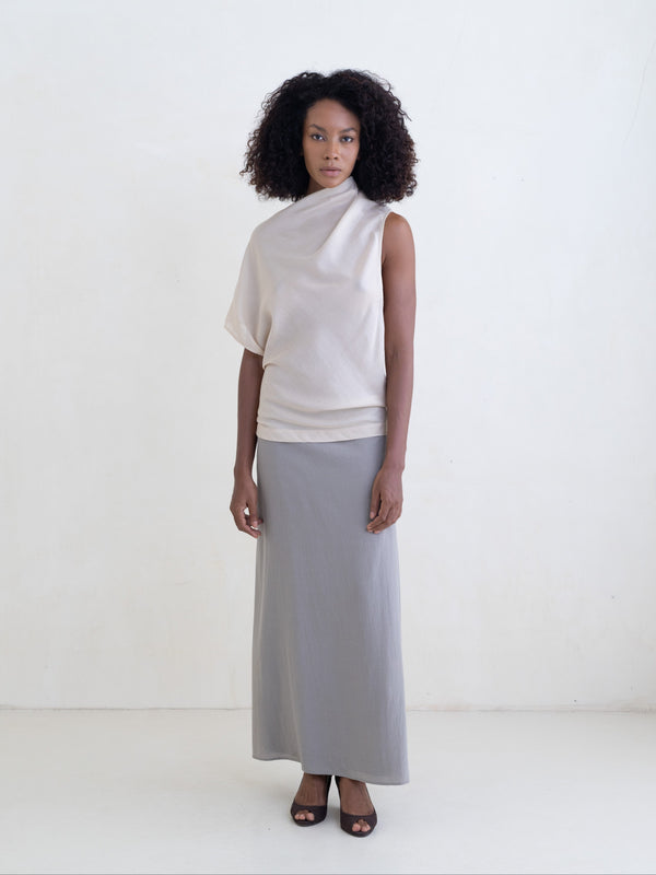 Woman wearing a cream draped one-shoulder top and a gray maxi skirt on a white background. 