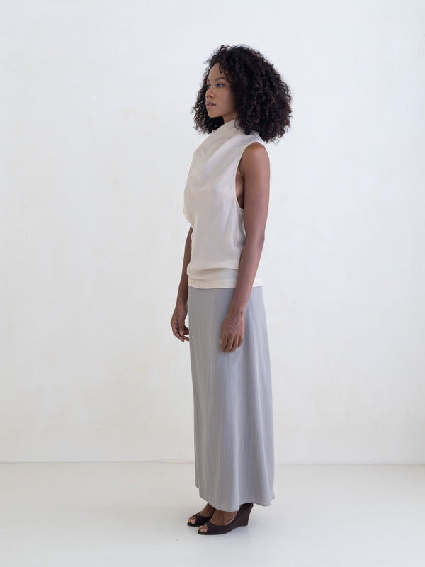 Woman wearing a cream draped one-shoulder top and a gray maxi skirt on a white background. 