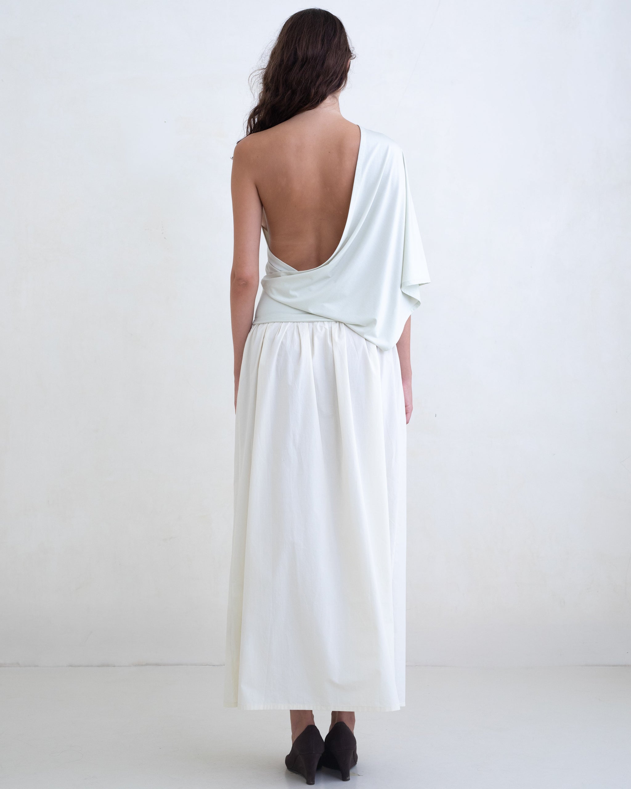 Woman wearing a light green one-shoulder top and a cream maxi skirt on a white background. 