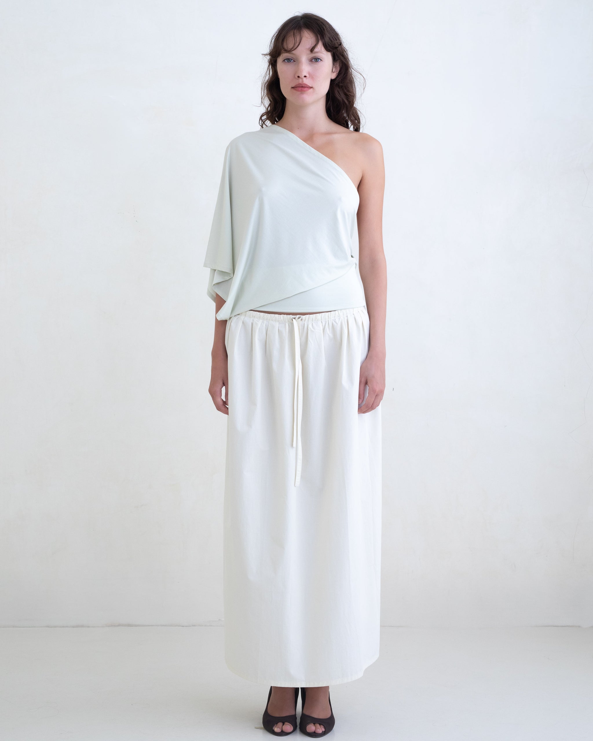 Woman wearing a light green one-shoulder top and a cream maxi skirt on a white background. 