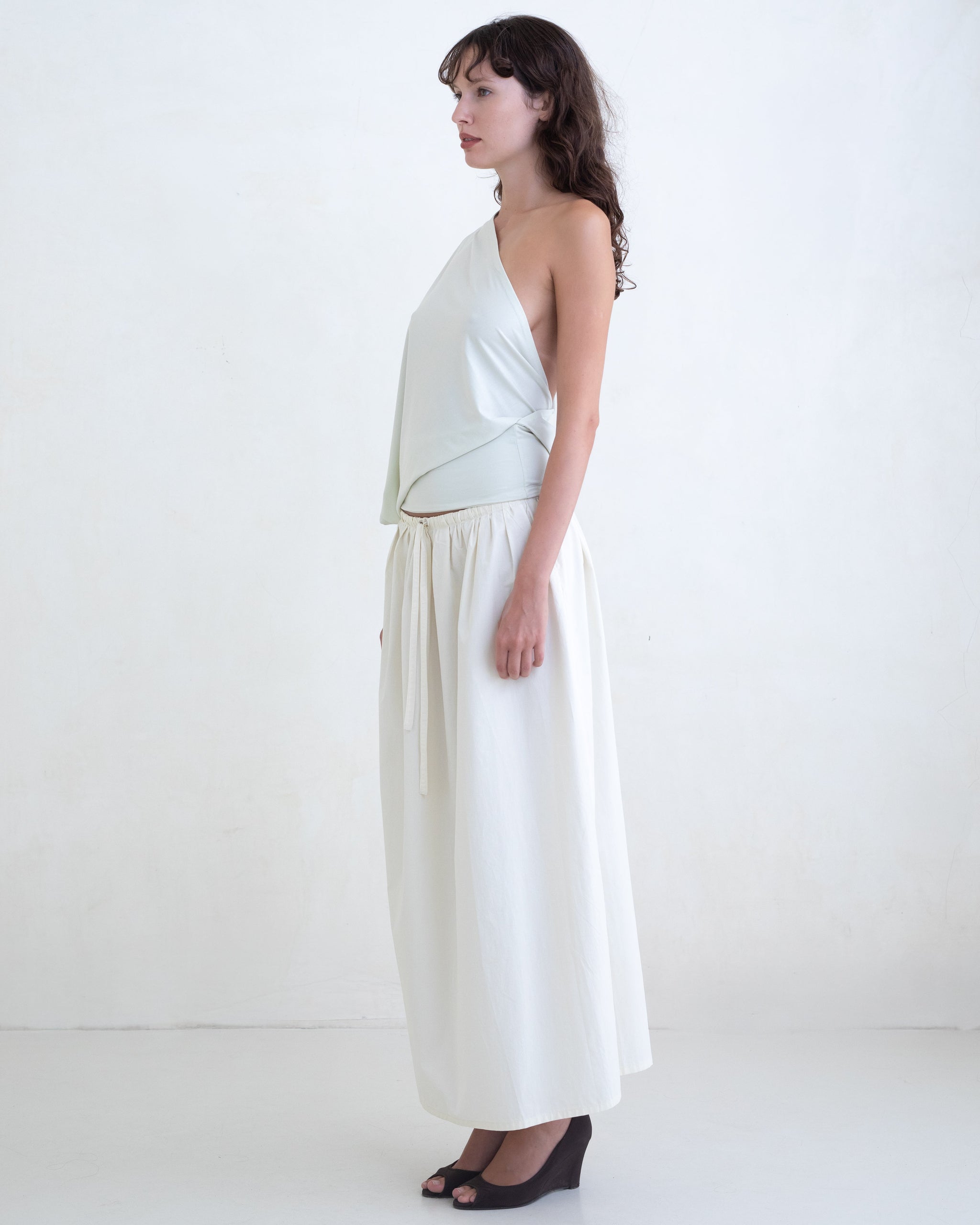 Woman wearing a light green one-shoulder top and a cream maxi skirt on a white background. 