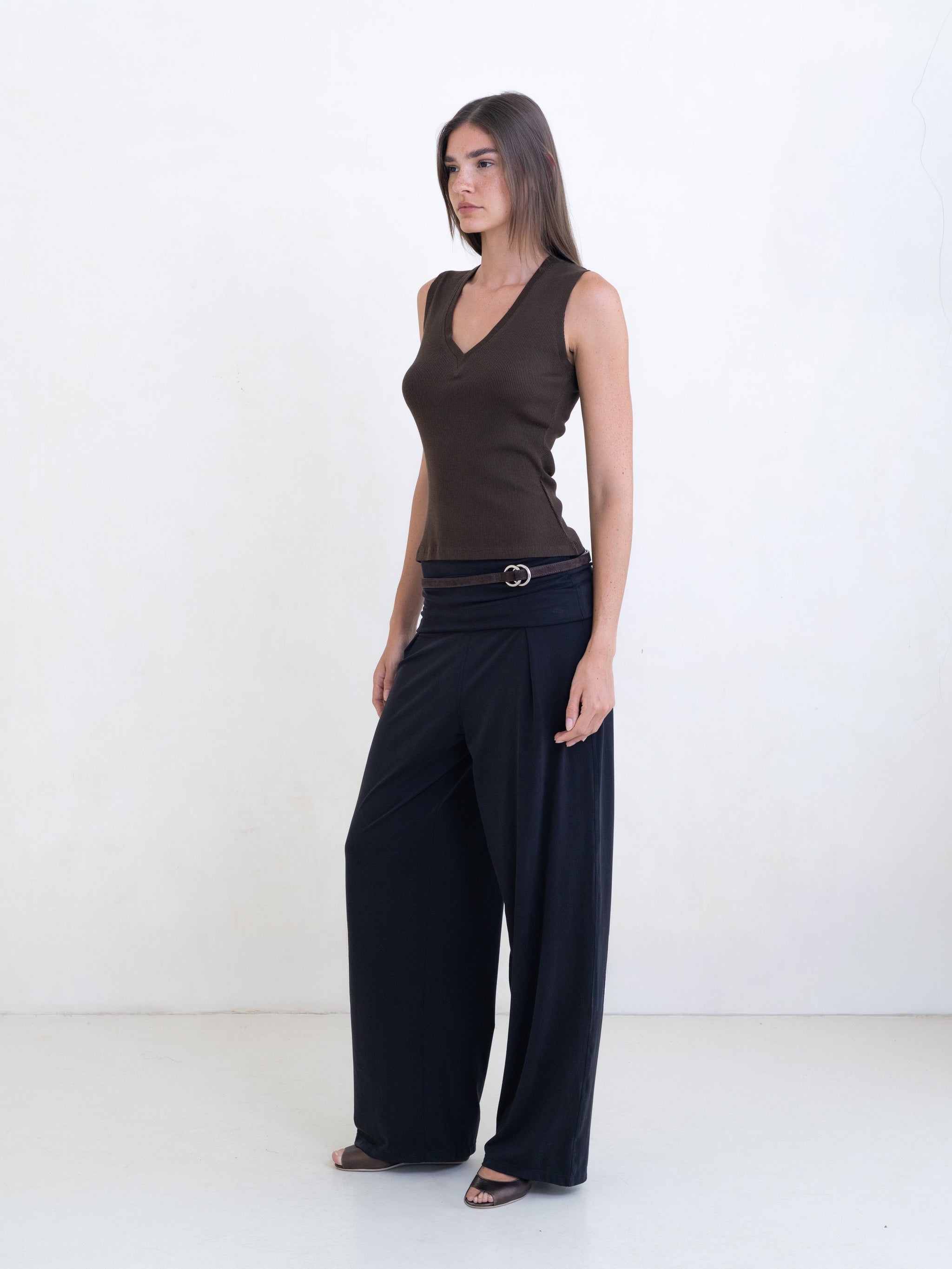 Woman wearing a brown sleeveless v-neck top, black pants with a brown leather belt on a white background. 