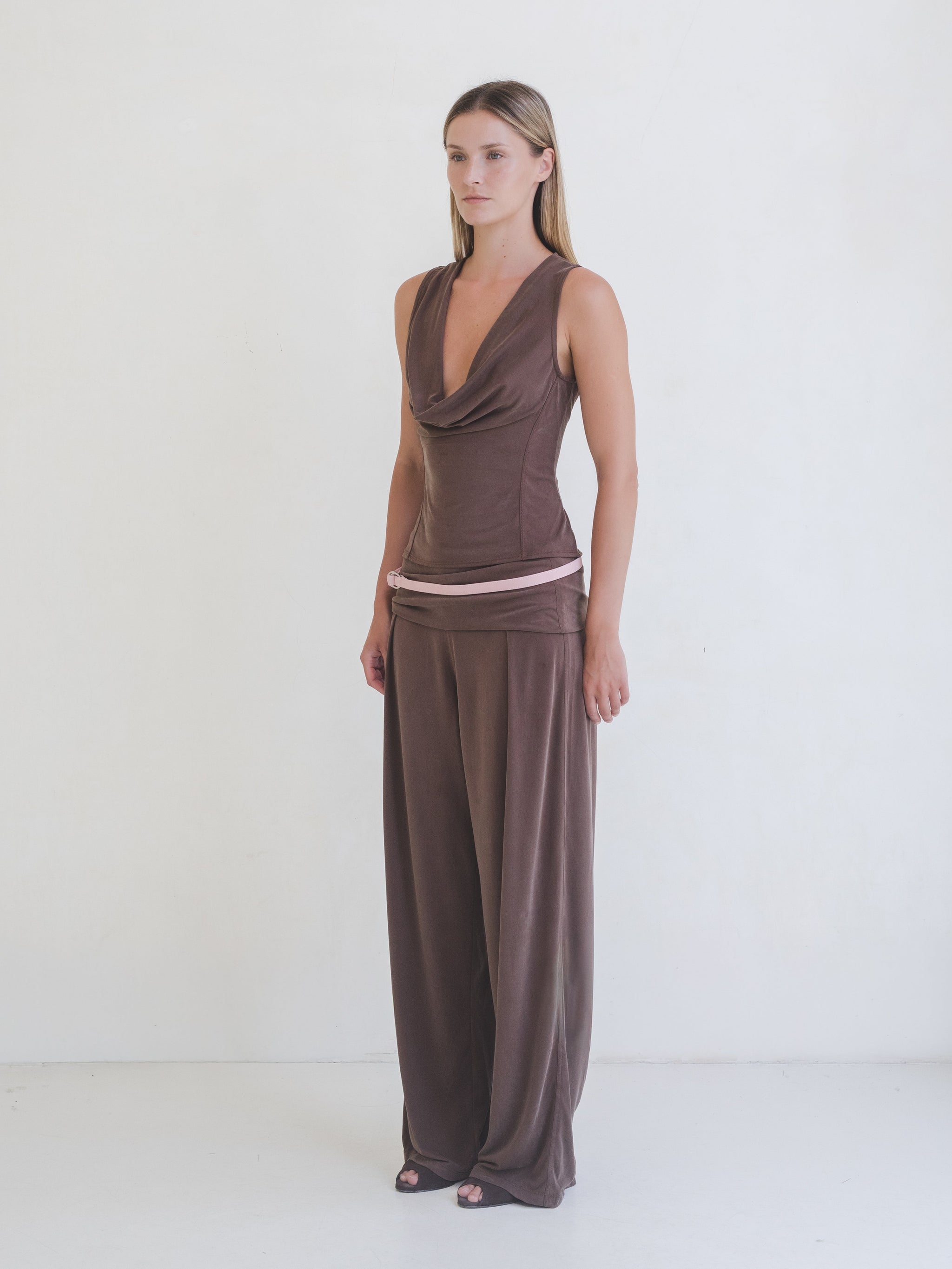 Woman wearing a brown sleeveless top, brown pants with a pink leather belt on a white background. 