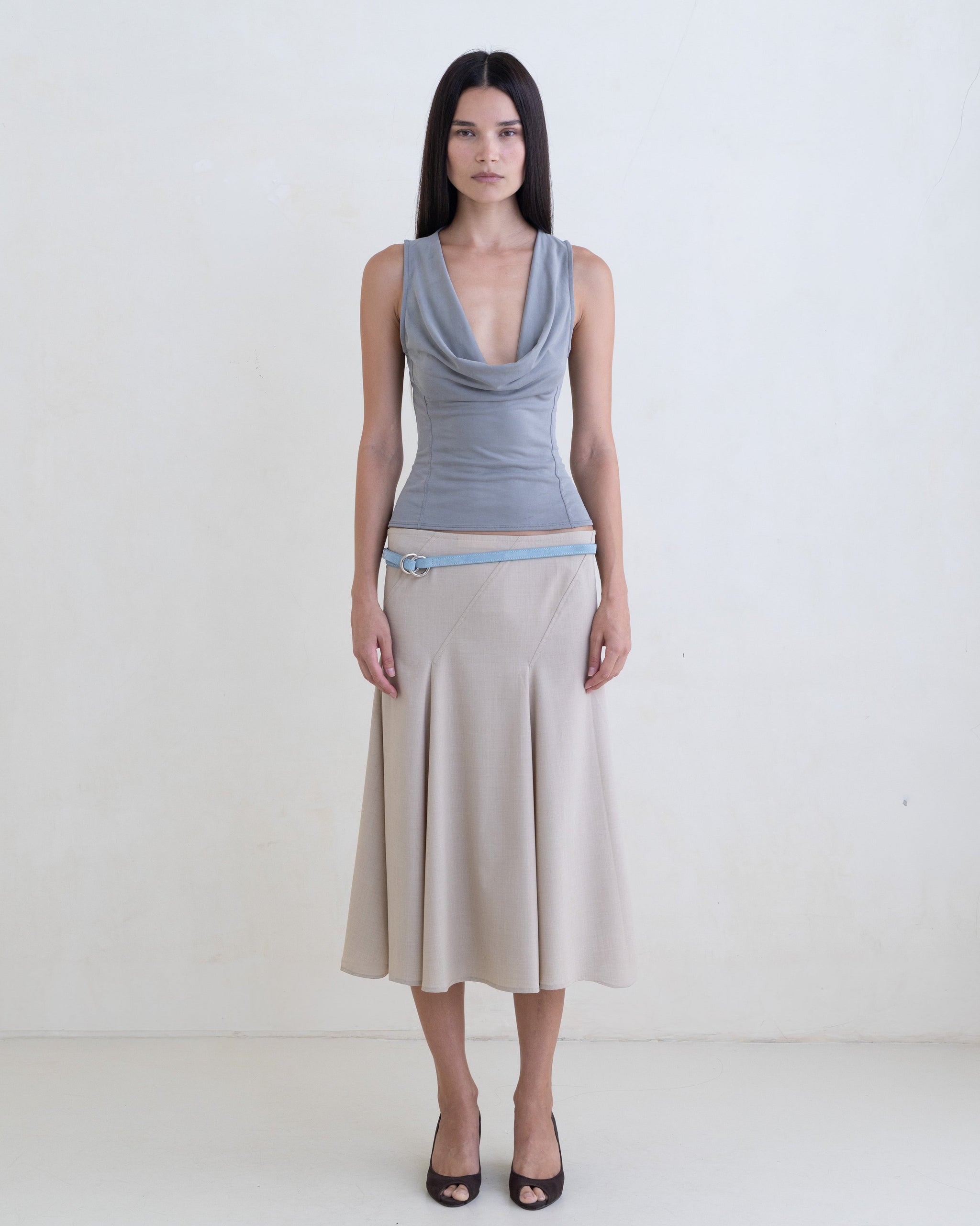 Woman wearing a gray sleeveless top, beige skirt with a light blue leather belt on a white background. 