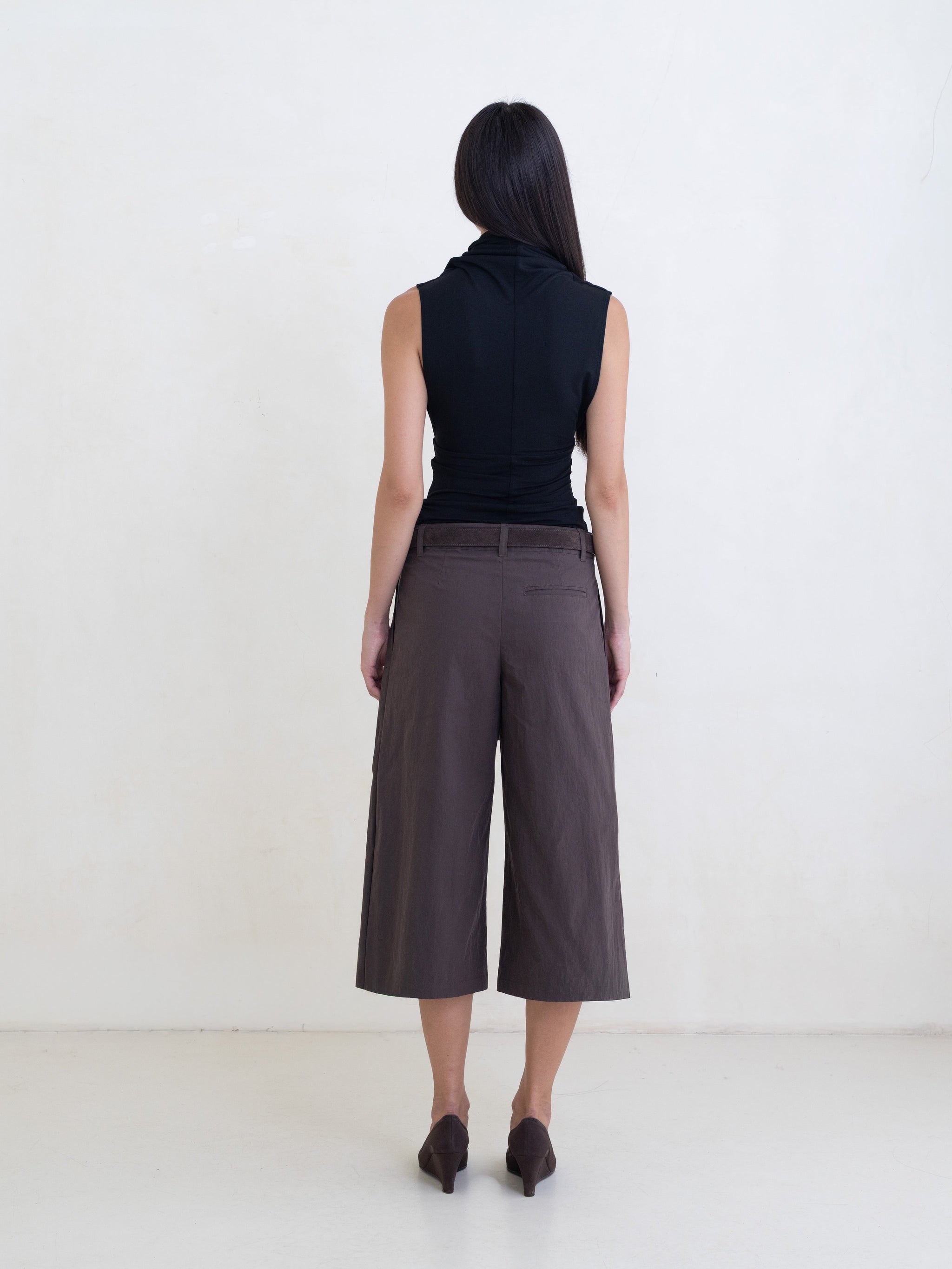 Woman wearing a black sleeveless top and brown wide-leg cropped trousers on a white background. 