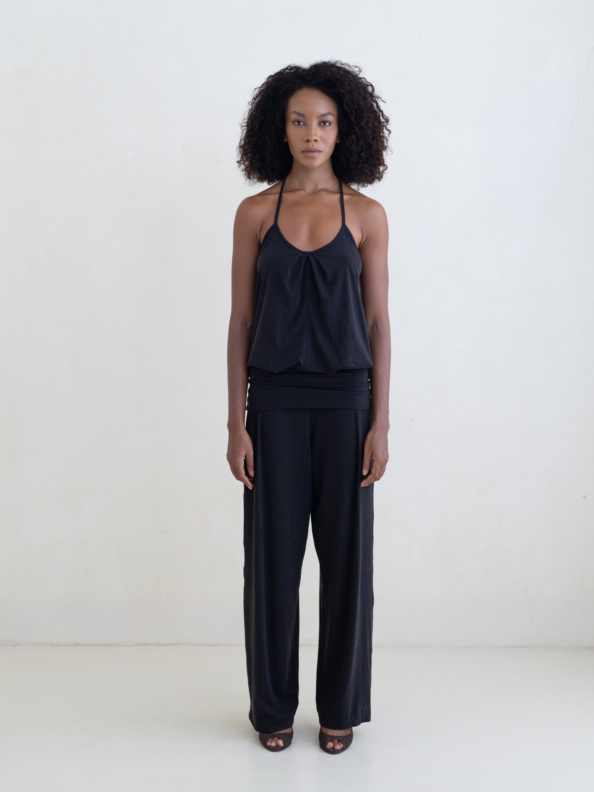 Woman wearing a black halter neck tank top and black straight leg pants on a white background. 
