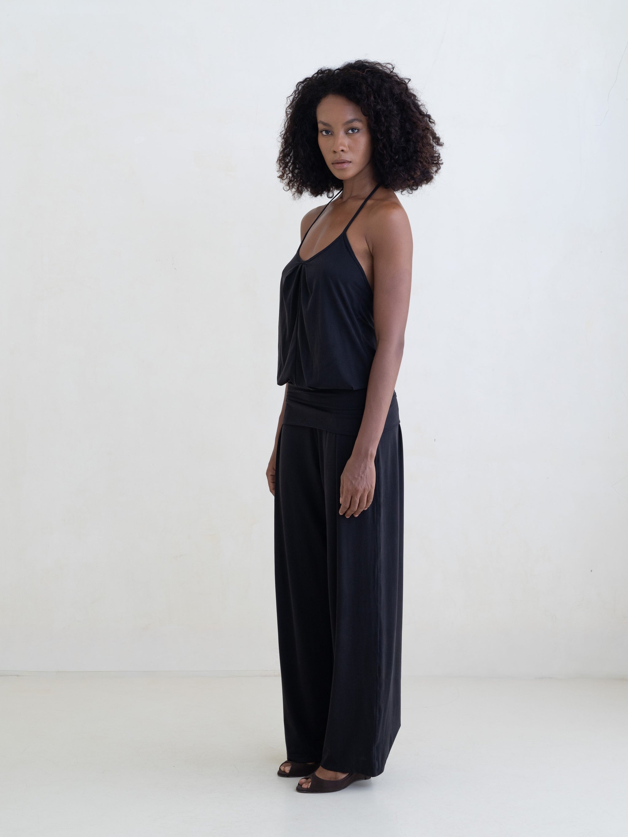 Woman wearing a black halter neck tank top and black straight leg pants on a white background. 