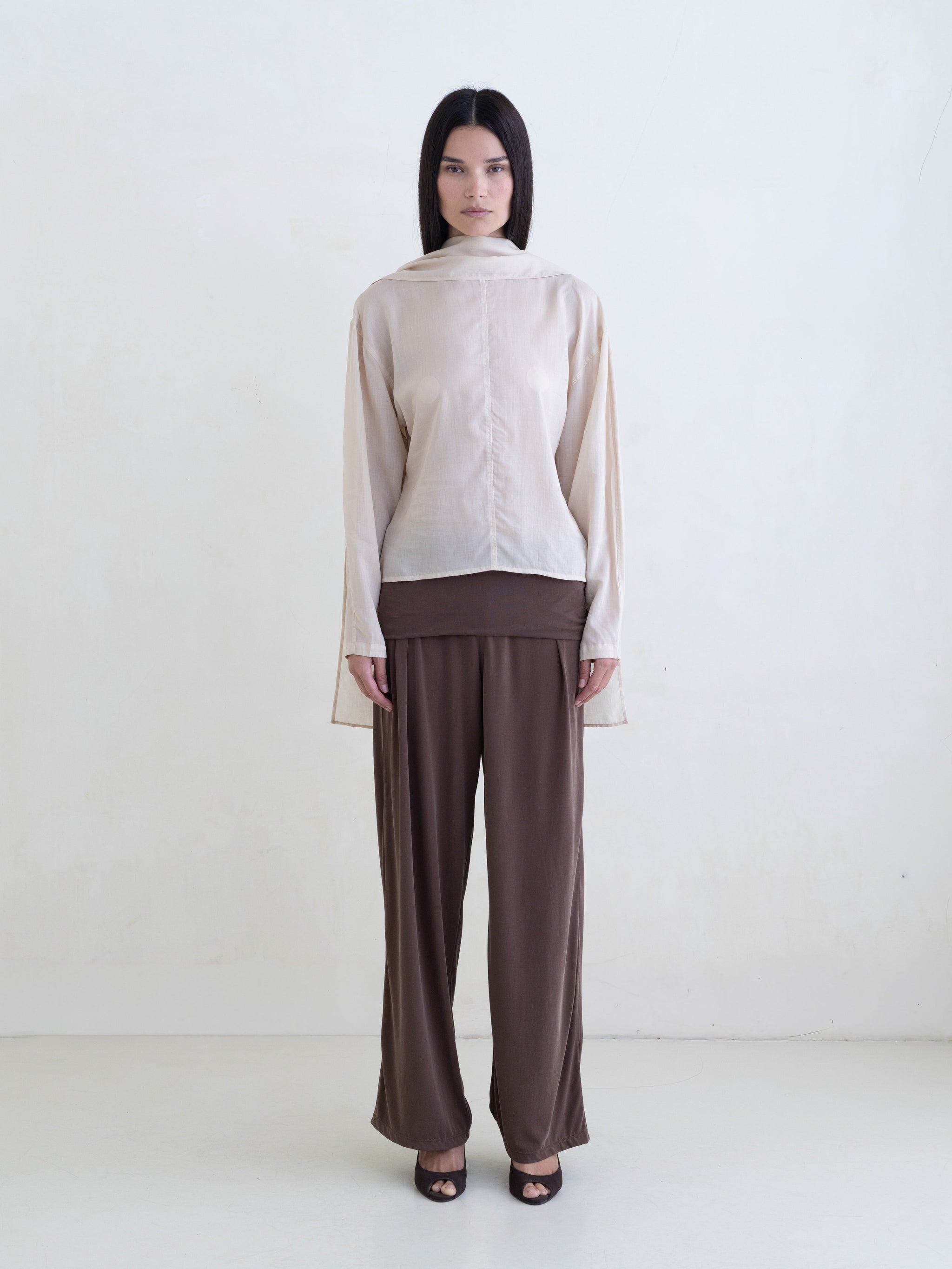 Woman wearing a cream long sleeve blouse and brown pants on a white background. 
