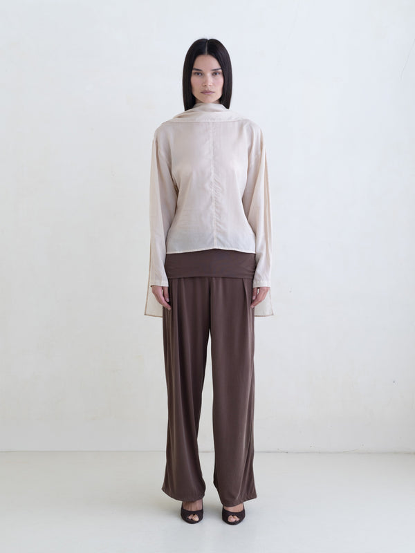 Woman wearing a cream long sleeve blouse and brown pants on a white background. 