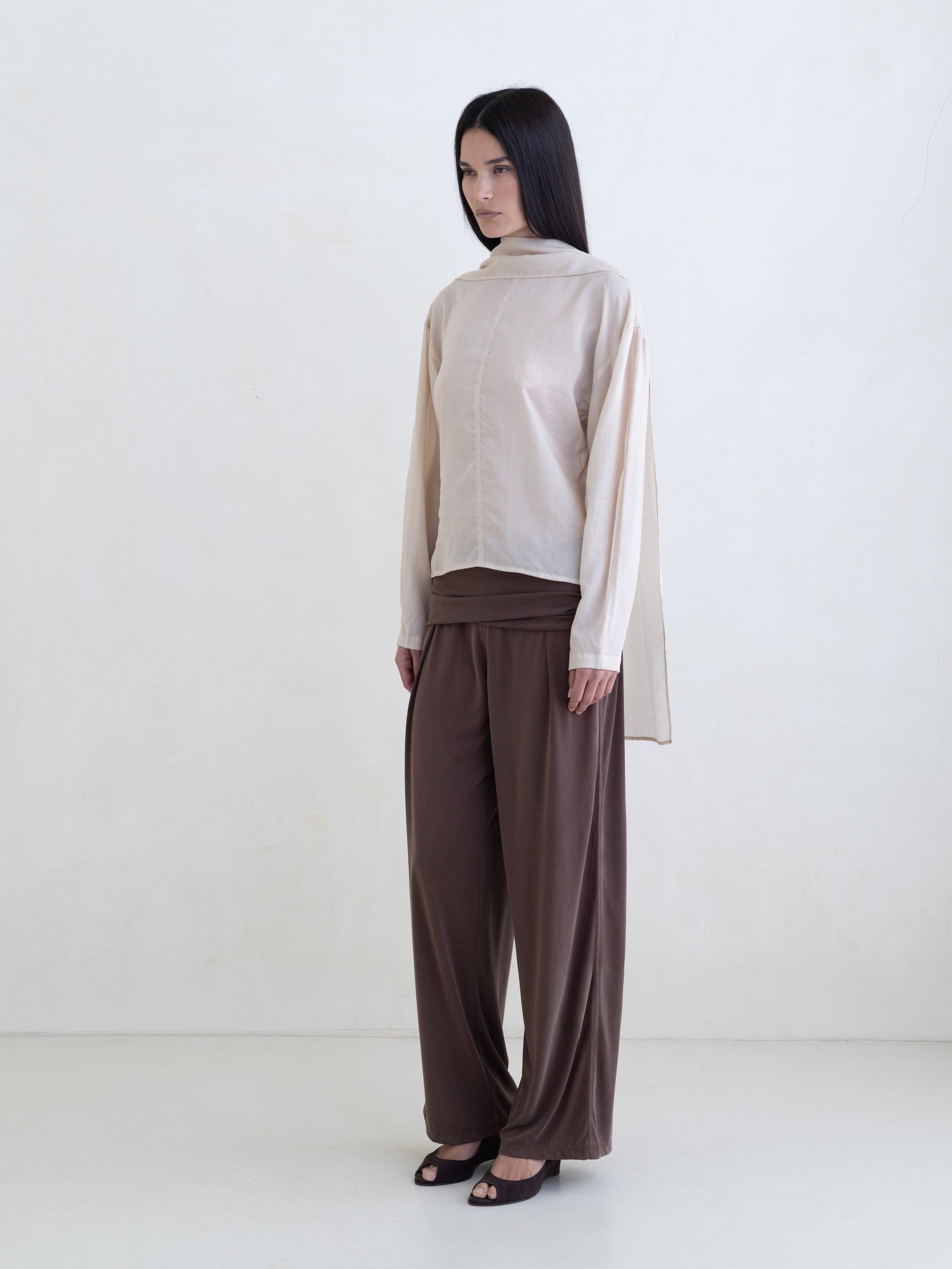 Woman wearing a cream long sleeve blouse and brown pants on a white background. 