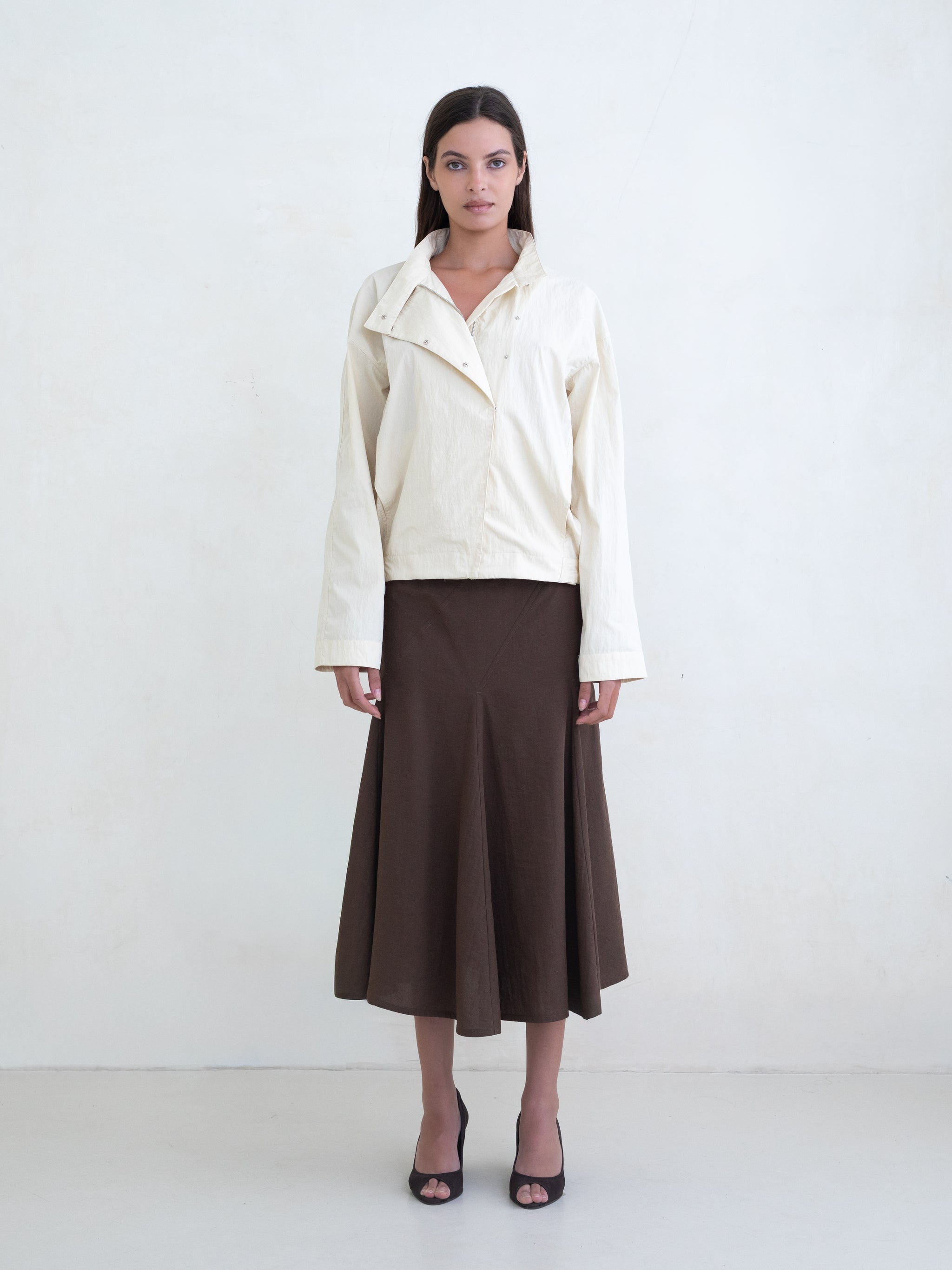 Woman wearing a cream asymmetric jacket and a brown midi skirt on a white background. 