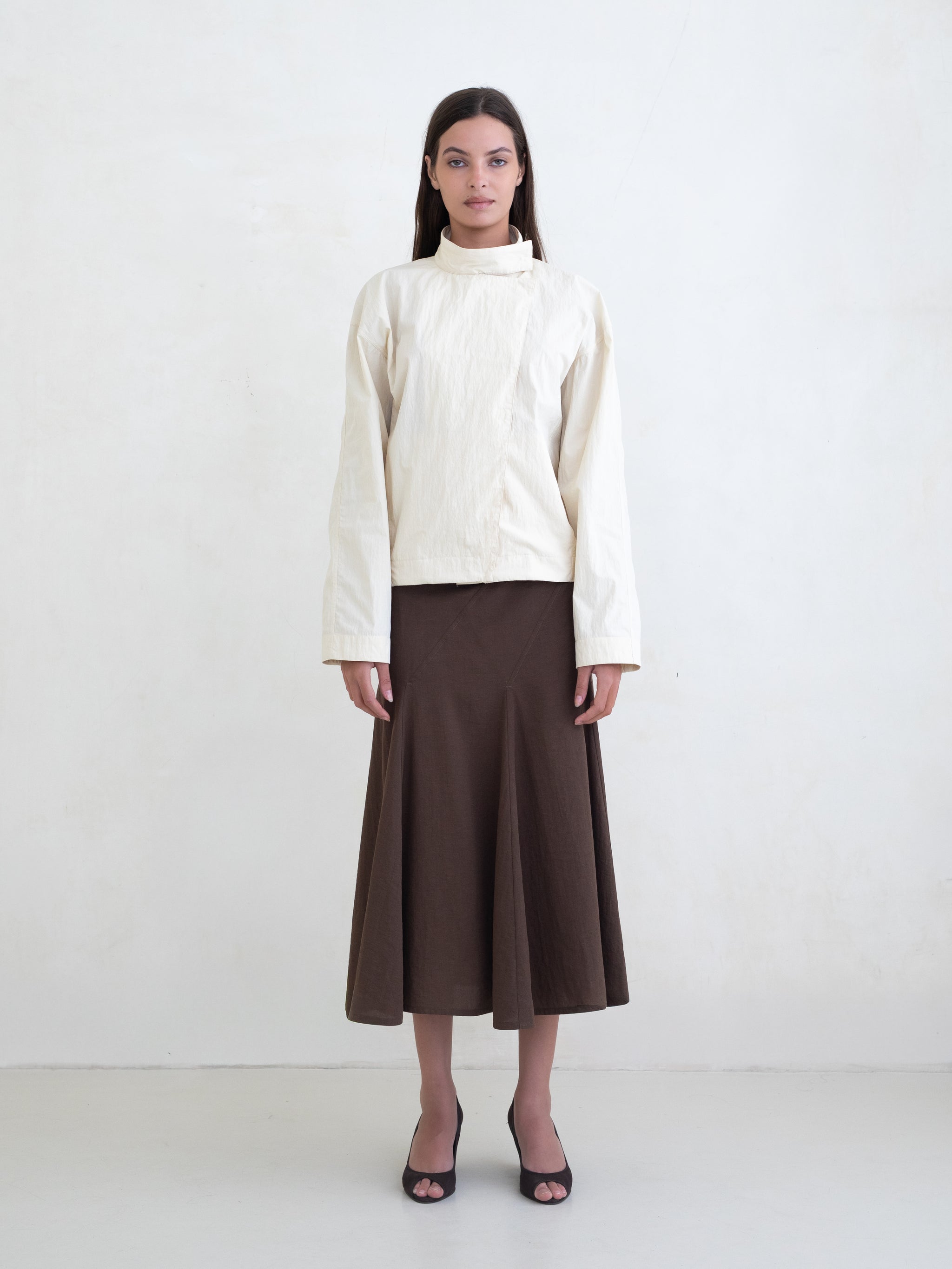 Woman wearing a cream asymmetric jacket and a brown midi skirt on a white background. 