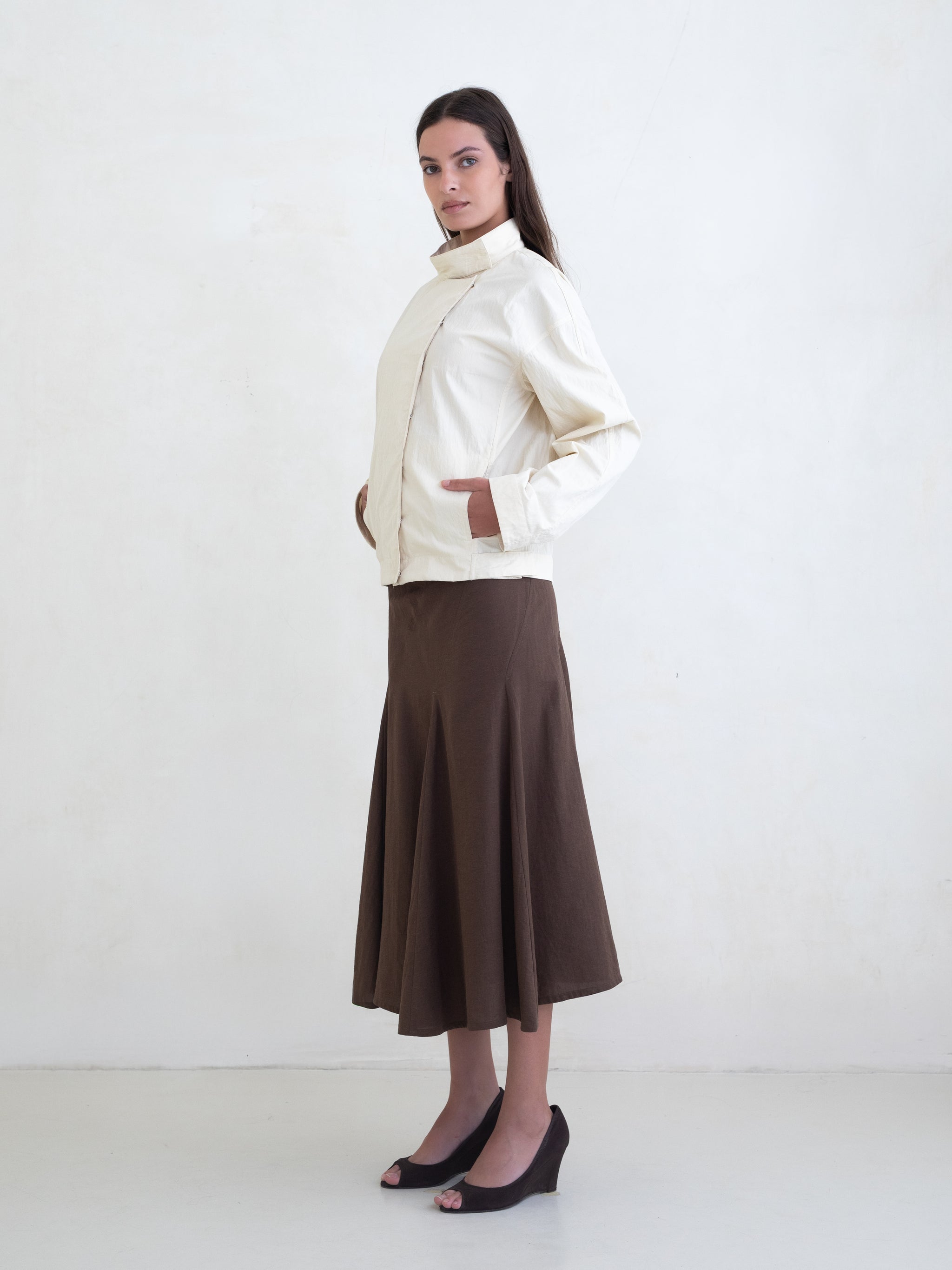 Woman wearing a cream asymmetric jacket and a brown midi skirt on a white background. 