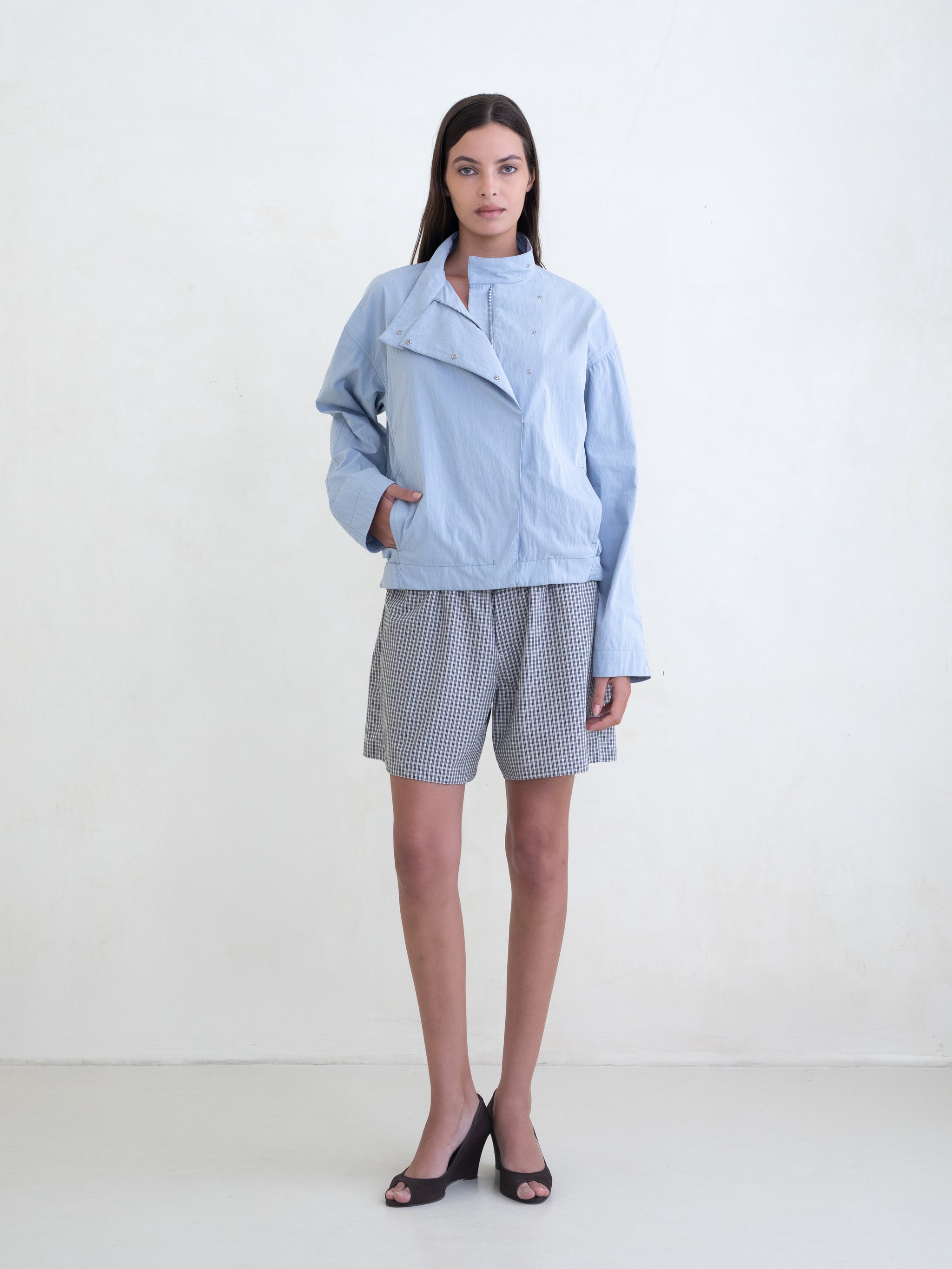 Woman wearing a light blue asymmetric jacket and a black plaid boxer shorts on a white background. 