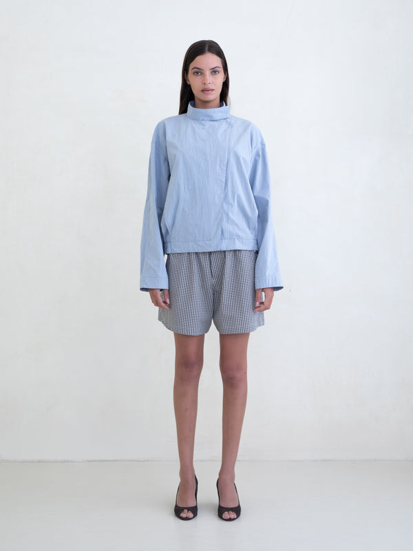Woman wearing a light blue asymmetric jacket and a black plaid boxer shorts on a white background. 