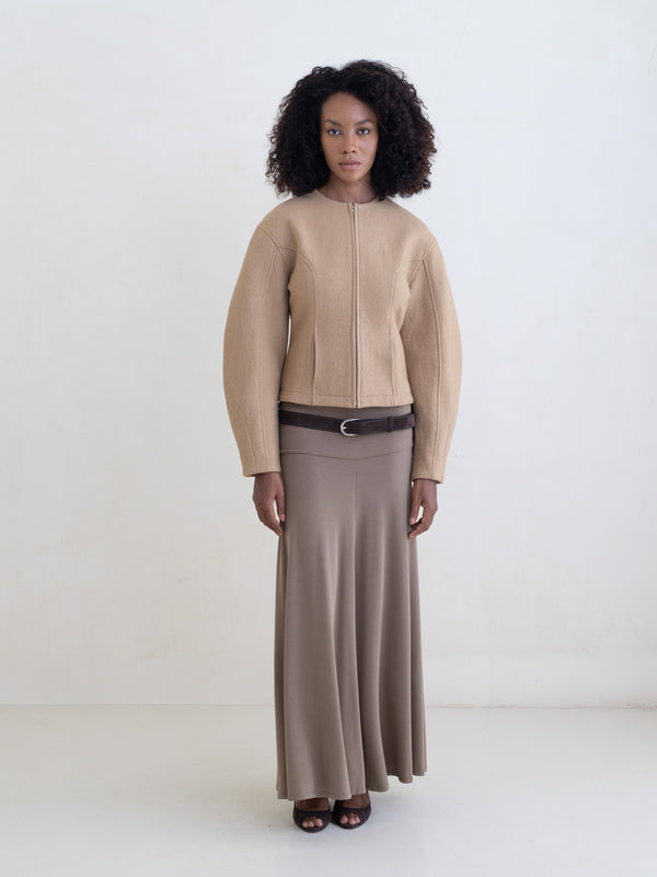 Woman wearing a beige zip-front jacket and a taupe maxi skirt on a white background. 
