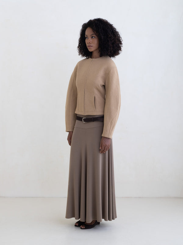 Woman wearing a beige zip-front jacket and a taupe maxi skirt on a white background. 